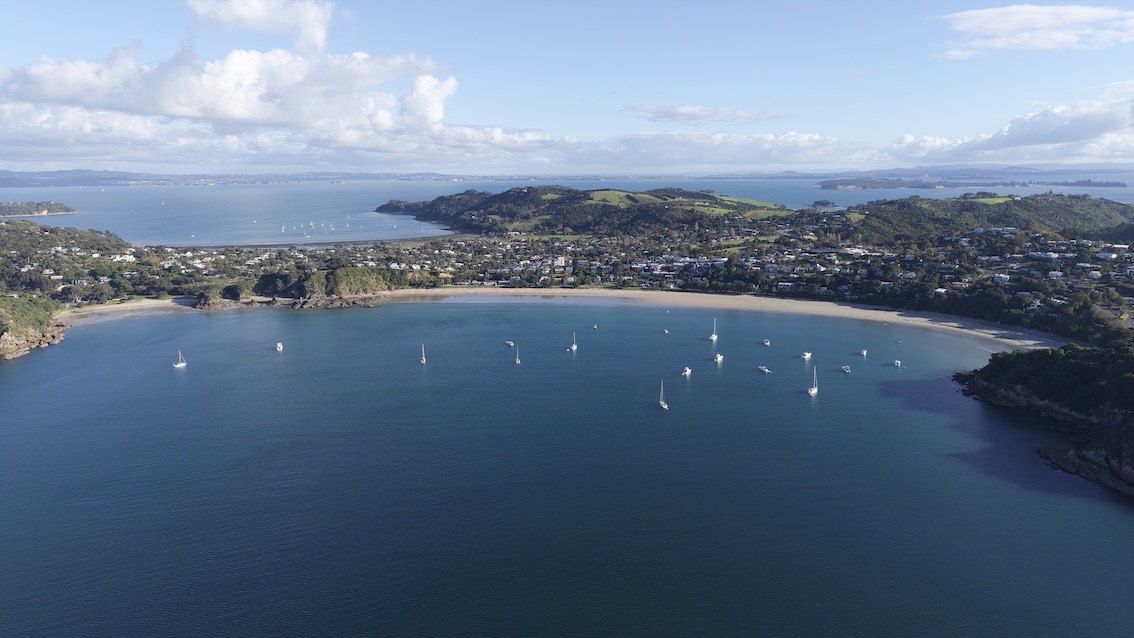 Oneroa Bay on Waiheke Island from the air: a curve of sand, yachts at anchor and green hills, with the Hauraki Gulf beyond